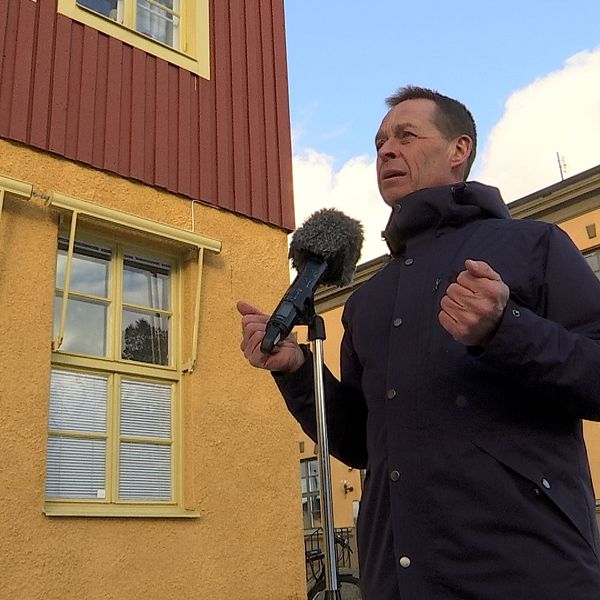 Man i blå jacka står vid ett mikrofonstativ. Tittar åt vänster. Bilden är tagen lite underifrån med två gulröda hus i bakgrunden.