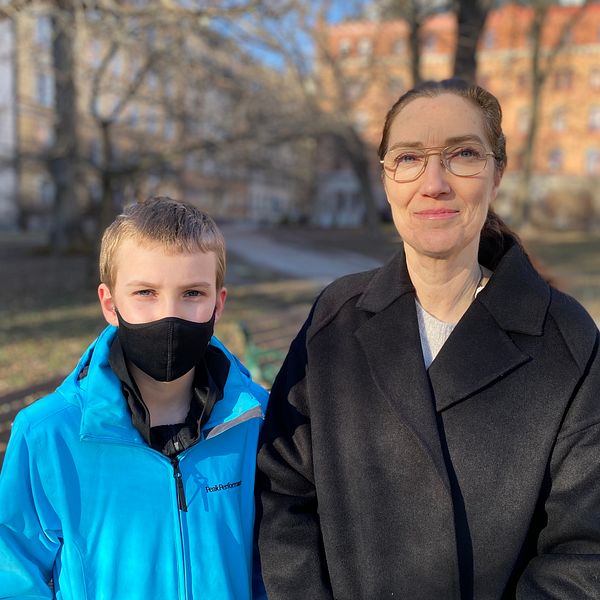 Linus har en blå jacka och ett svart munskydd på sig. Brevid står hans mamma Sarah, hon har en svart jacka och glasögon. De är i fokus medan bakgrunden är lite suddig.