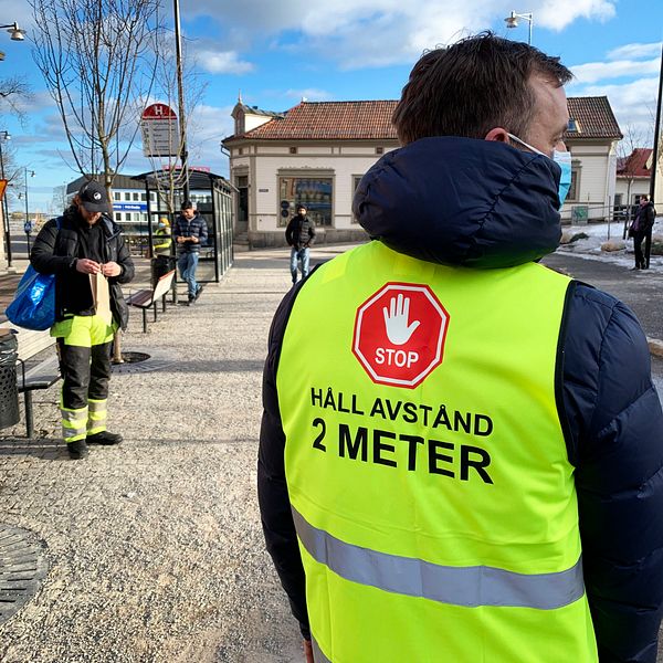 Man i reflexväst på en busshållplats. Han står med ryggen mot kameran. På ryggen står det ”STOP HÅLL AVSTÅND 2 METER”. Han bär munskydd.