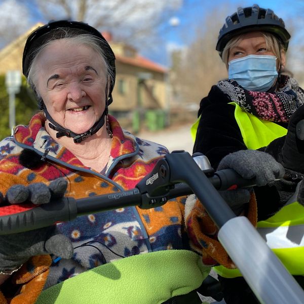 Halvbild på 90-åriga Kerstin Rause och Rose Andersson som jobbar på Rosengårdens äldreboende. Båda har hjälm på sig och har händerna på varsitt styre. Rose har munskydd och en gul reflexväst på sig. Kerstin ler och kollar in i kameran, hon har på sig en fleecetröja i rött, orange, blått och vitt.
