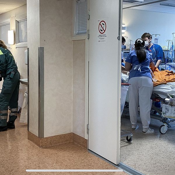 personal från ambulanshelikopter rullar in en brits i korridoren medan intensivvårdspersonal ses vårda en coronapatient i ett rum