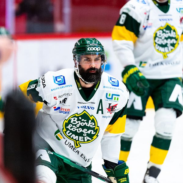 En man i grön ishockeyhjälm och tätt skägg står på knä på en isrink med fokuserad blick.