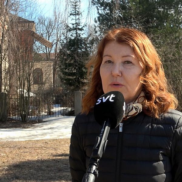 en kvinna intervjuas utomhus, träd och äldre byggnader i bakgrunden, snöfläckar