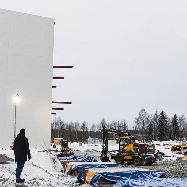 Två traktorer som jobbar med att bygga upp en ny fabrik. En stor vit vägg och en människa framför.