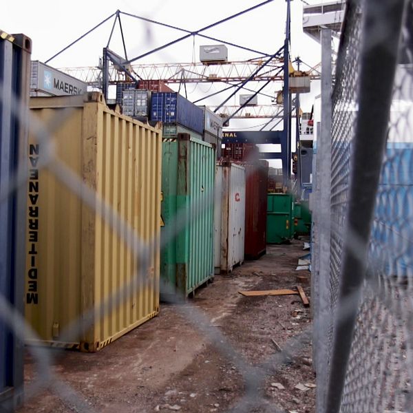 containrar på rad ses genom ett staket i hamnområdet, kranar i bakgrunden