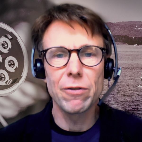 En person med hörlurar står framför en bakgrund där en båt syns som åker på vattnet, med en naturskön utsikt i bakgrunden. Till vänster i bilden syns en laddkontakt för elfordon.