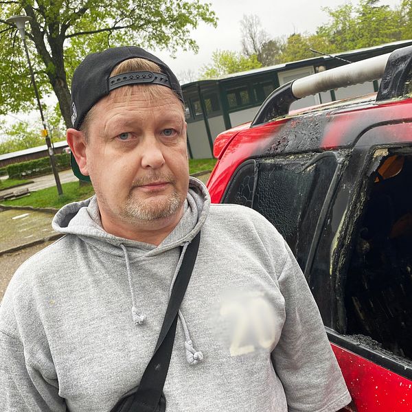 Lars står intill sin utrbrända bil på en parkeringsplats.