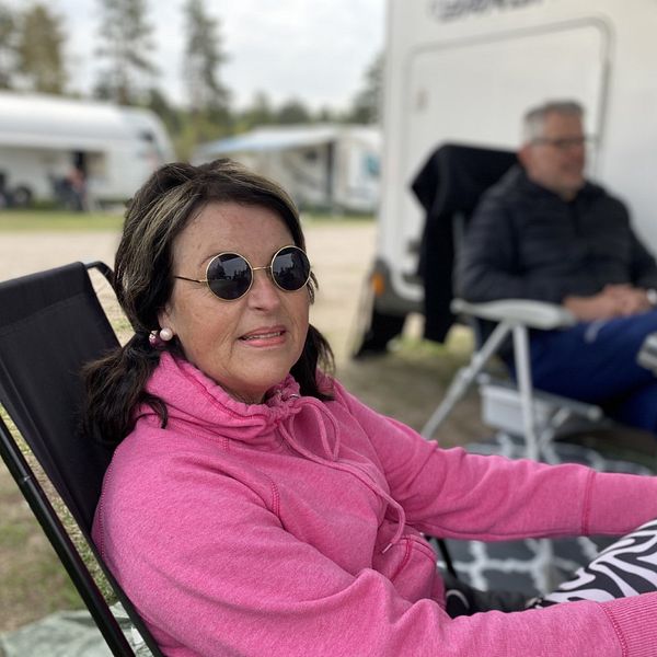 en medlelålders kvinna i solglasögon och rosa luvtröja sitter i en campingstol utanför husvagn