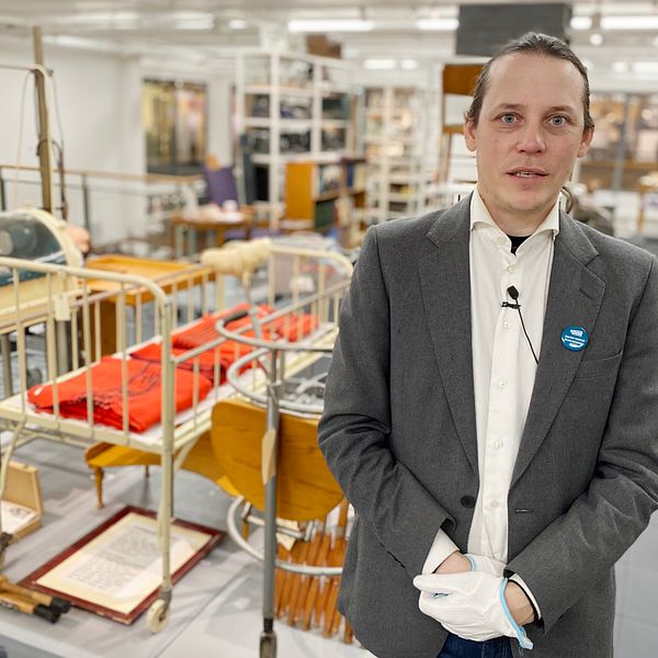 Jean-Pierre Salkvist står intill några sjukhusföremål från 1900-talet, bland annat en säng, ett par kryckor och en docka.