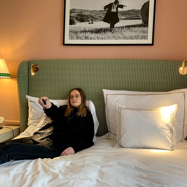 Reporter lutar sig tillbaka i hotellsängen och håller en fjärrkontroll i handen. Till vänster i bild syns en lampa, väggarna är i persikofärg och över sängen hänger en tavla.