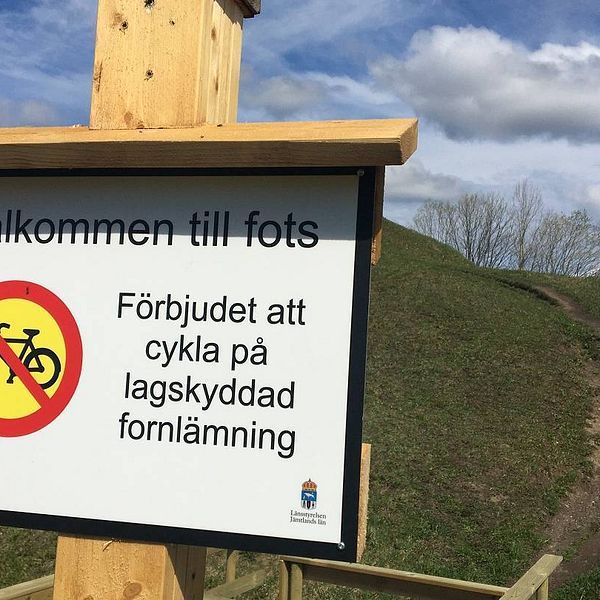 Skylt med texten ”Välkommen till fots. Förbjudet att cykla på lagskyddad fornlämning”. I bakgrunden en rejäl stig uppför en kulle.