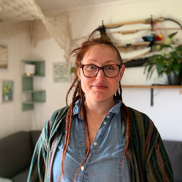 Tanja Östberg har röda dreadlocks och glasögon. Tanja har en jeanblus och en cardigan på sig. Hon står i sitt vardagsrum framför soffan och poserar. I bakgrunden hänger några pilbågar på väggen.