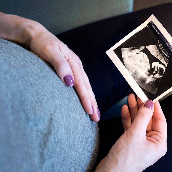 närbild ner mot en kvinnas gravidmage, ena handen på magen, andra håller en ultraljudsbild