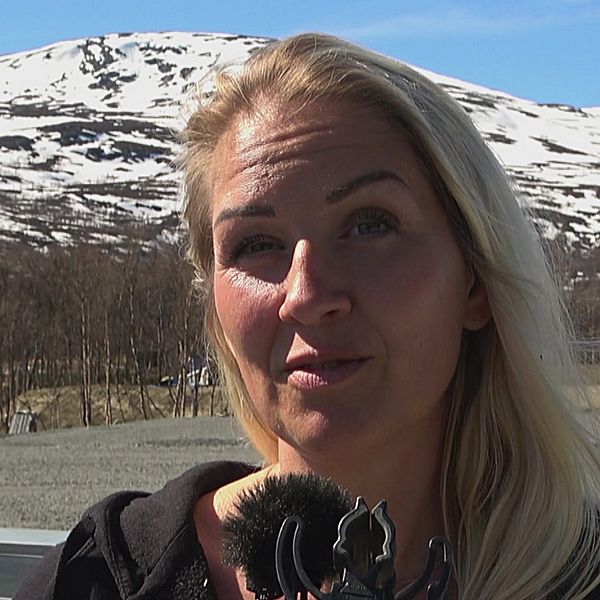 kvinna som intervjuas med snöfläckiga fjäll i bakgrunden, husvagn skymtar