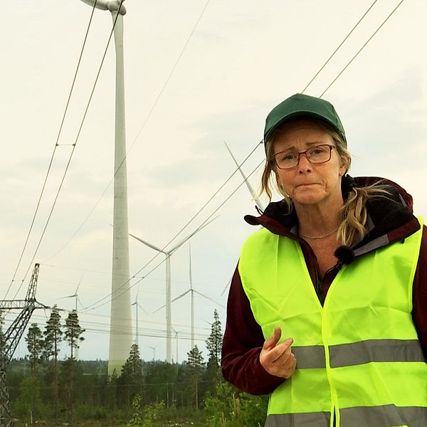 En kvinna i gul varselväst med vindkraftverk i bakgrunden.