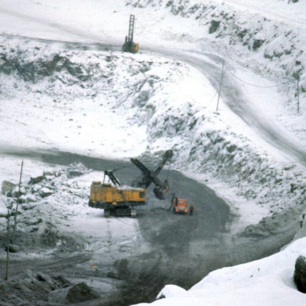 en gruvmaskin, en bil och en människa syns nere i en jättelik grusgrop med en väg ner, tunt snötäcke