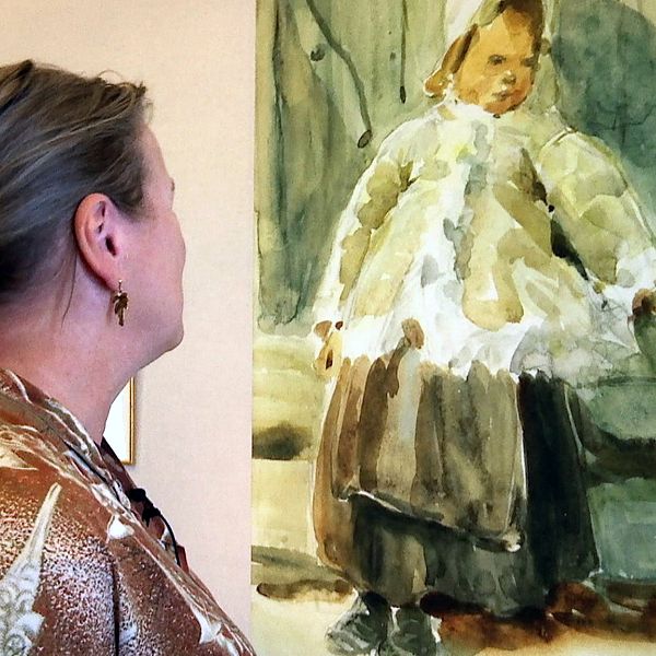 kvinna ses snett bakirån, tittar på akvarellmålning av ett barn