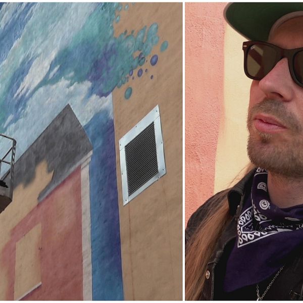 Till vänster en skylift med en konstnär som håller på att göra en muralmålning på en husvägg. Till höger en man med svarta glasögon och vit keps.