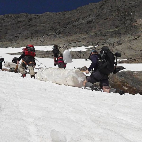 flera personer med ryggsäckar drar två stora plastade rullar uppför en brant sluttning med snö och klippor