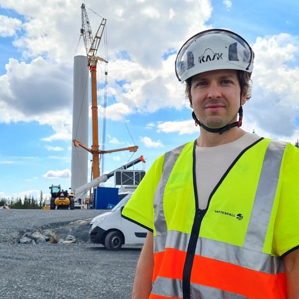 En man med gul och orange reflexväst och vit hjälm står framför byggnationerna av ett vindkraftverk.