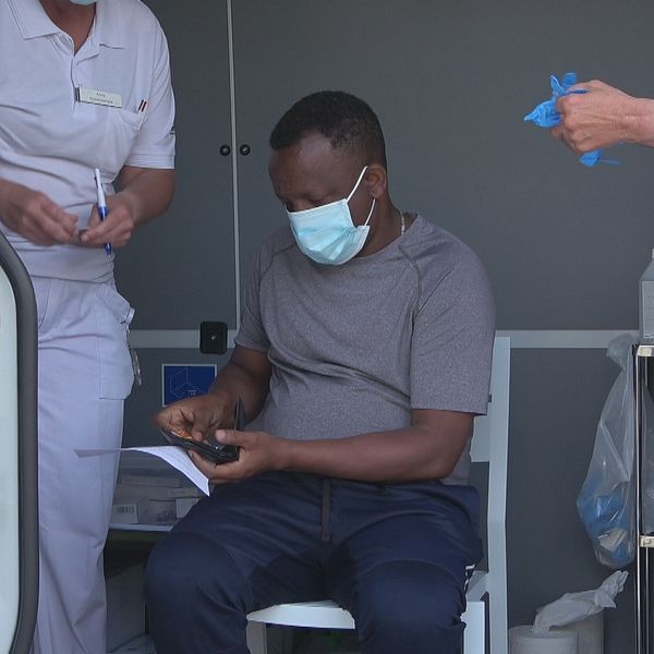 En man sitter på en stol och bär munskydd och grå t-shirt väntar på att få sitt vaccin. En sköterska klädd i vitt till vänster och en sköterska klädd i vitt till höger om mannen.