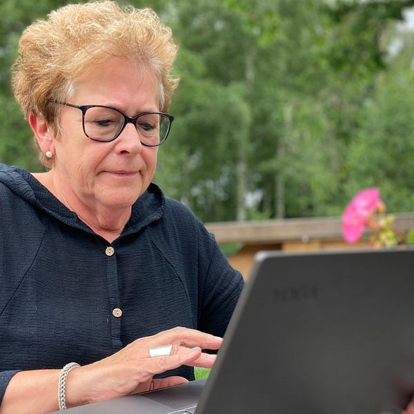 Bild på en kvinna i 60-årsåldern som sitter framför en svart laptop. Hon sitter utomhus på en veranda, i bakgrunden skymtar grönska.
