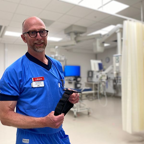 Läkaren Jacob Broms visar upp det avsnörande förband som är bra att använda för att stoppa en blödning. Han har blå kläder, bakgrunden är suddig men han står i ett akutrum på sjukhuset.
