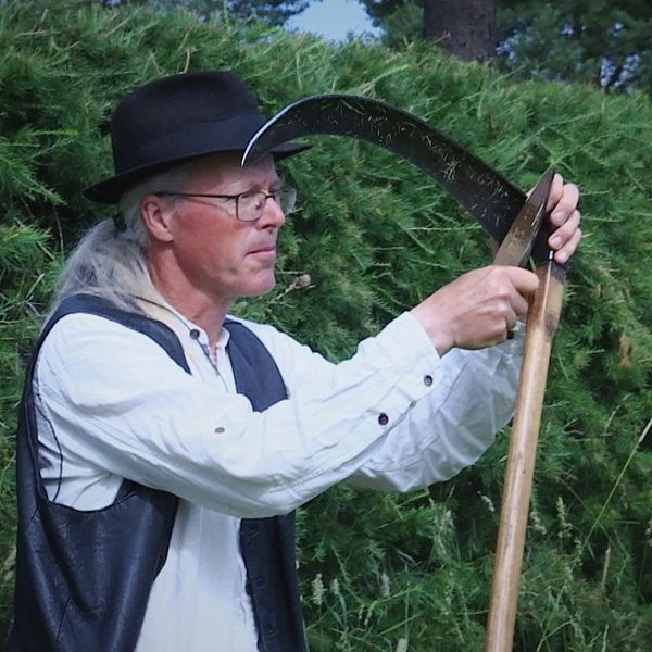 en man med glasögon och lång grånat hår, i gammaldags hatt, skjorta och väst, vässar en lie