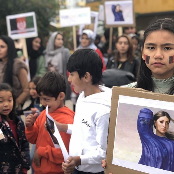 flicka med afghanska flaggan målad på kinderna, håller ett plakat med en bild, flera barn och personer med plakat i bakgrunden