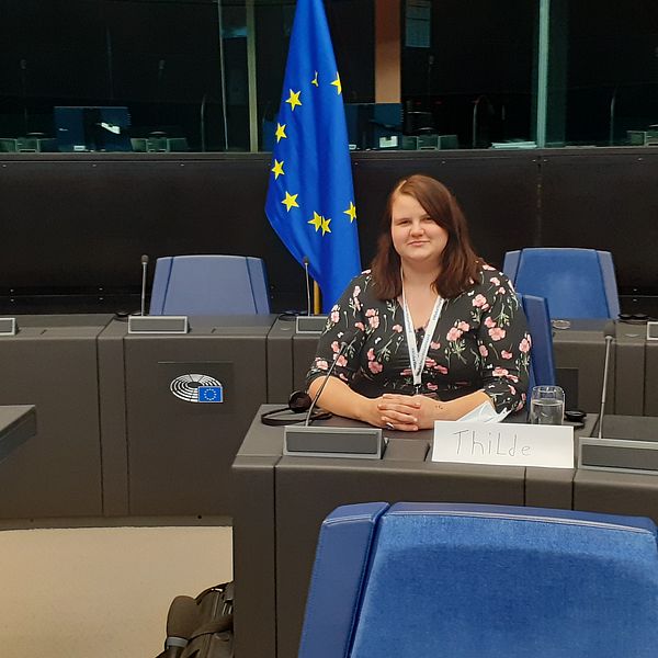 Thilde sitter vid ett bord. bakom sig har hon EU:s flagga.
