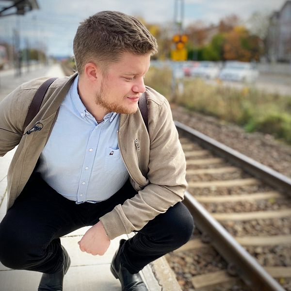 Mattias Winsa, i blå skjorta och beige jacka, sitter på huk på perrongen och tittar ner mot tågspåret.