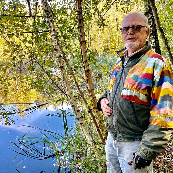 En man i färgglad jacka står framför en liten damm med spegelblankt vatten
