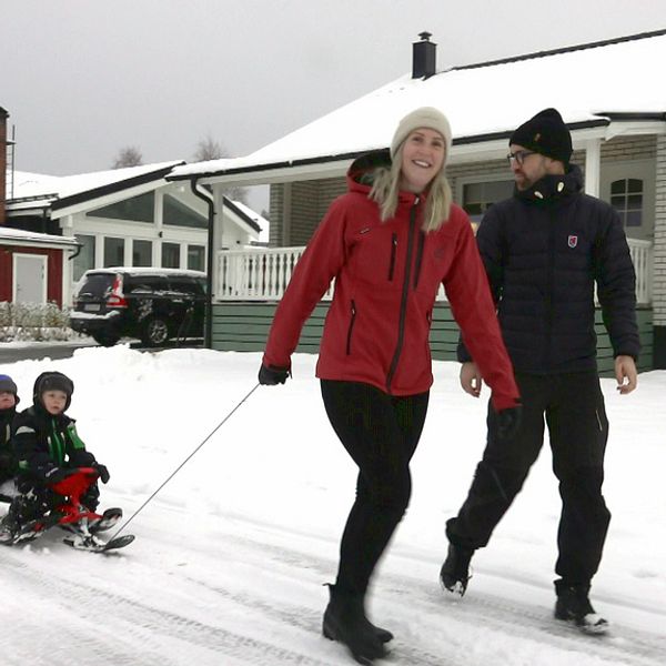 En man och en kvinna går på en snöig väg. Kvinnan drar två små barn på en bob. I bakgrunden syns villor.