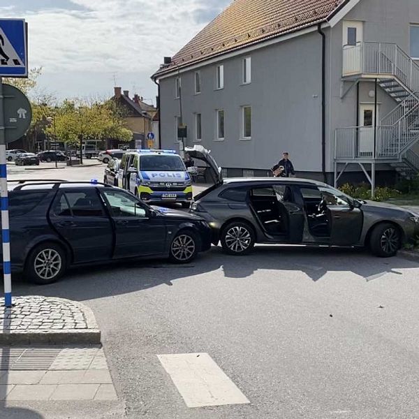 En svart civil polisbil har kört in i en grå bil och tvingat den till kanten strax intill ett övergångsställe. Bildörrarna och bakluckan står öppna. I bakgrunden syns en större polisbil.