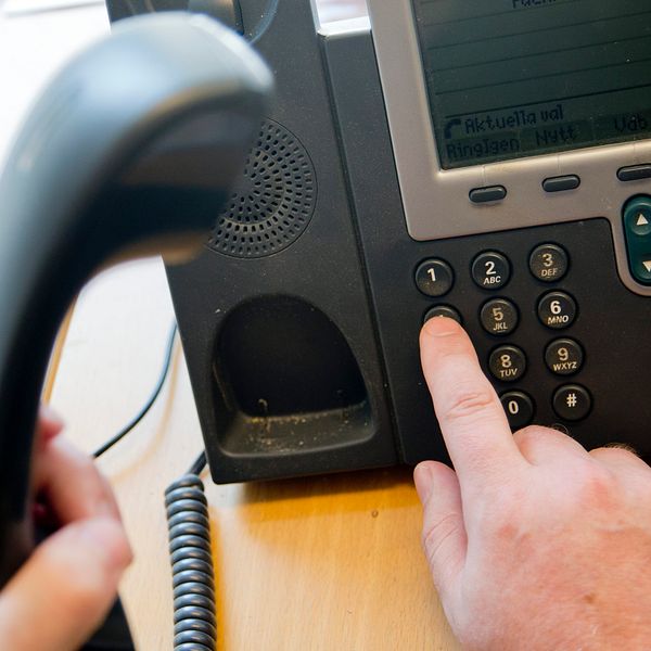 En person lyfter en telefonlur och har lagt ett finger på nummerpanelen för att slå ett nummer.