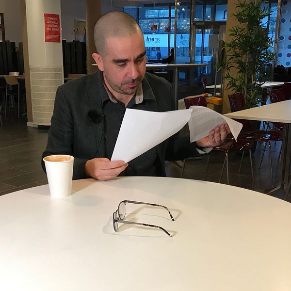 Person läser dokument vid cafébord; glasögon och kaffemugg på bordet.