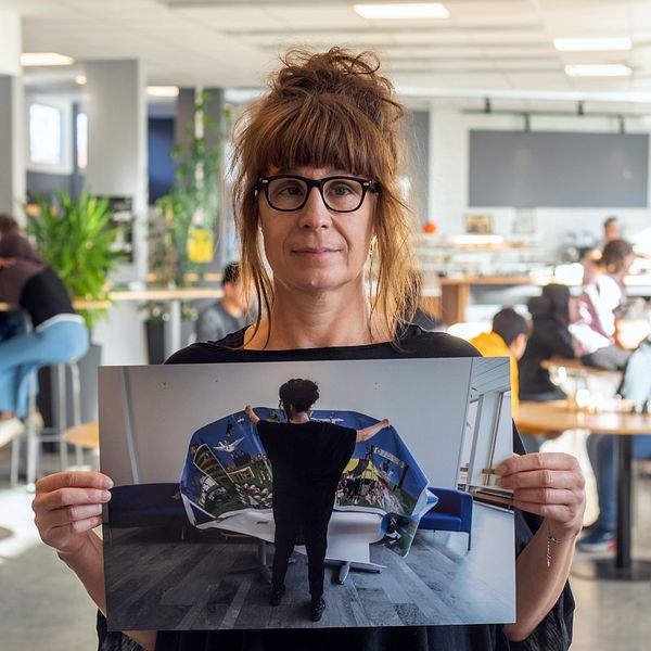 Kvinna i skolmiljö håller upp bild på en kvinna om håller i en affisch