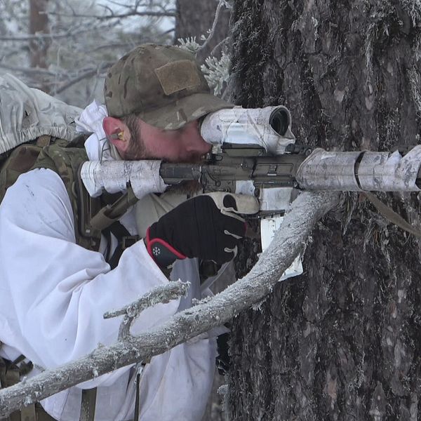 Jonas Dahl, en soldat i vinterkamouflage med stor ryggsäck siktar med sitt vapen intill ett träd