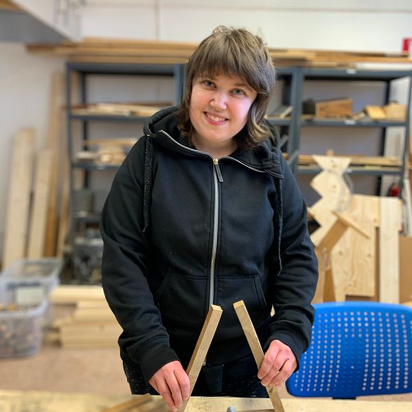 Helena står i en snickarverkstad och ler stort mot kameran.