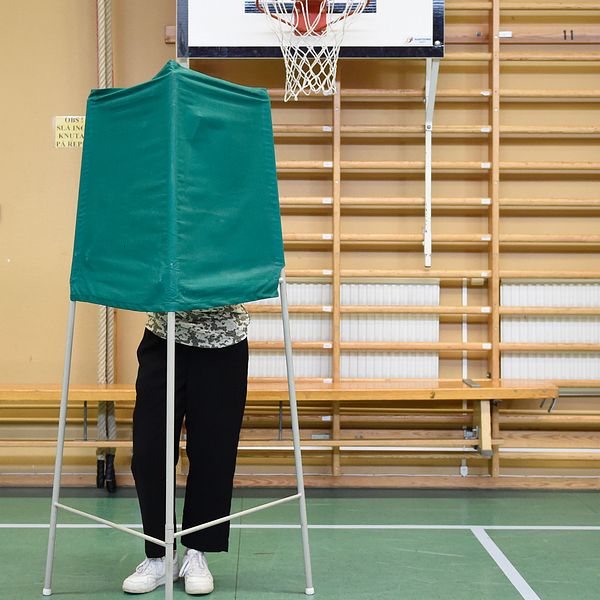 Bild från en vallokal (gymnastikhall). En person står bakom ett grönt valbås