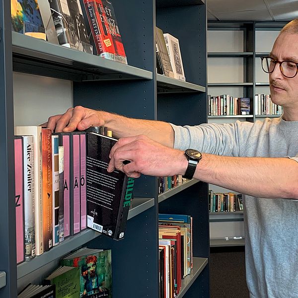 Man i glasögon bläddrar bland böcker i bokhylla på bibliotek