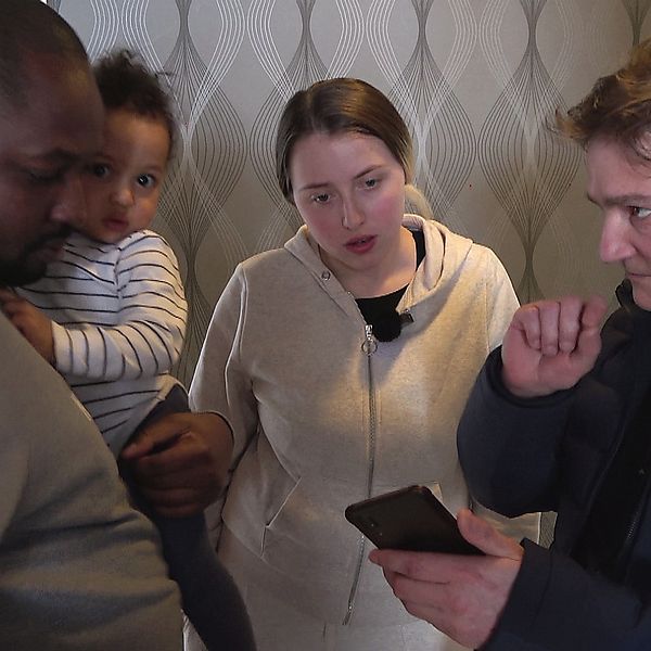 en man med ett litet barn på armen och en kvinna, som pratar med en medelålders man med mobiltelefon
