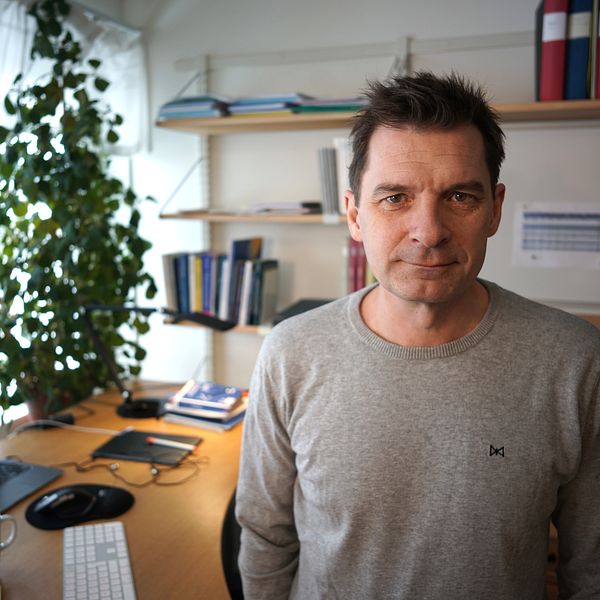 En man i grå stickad tröja tittar rakt in i kameran ståendes i sitt kontor på Umeå universitet