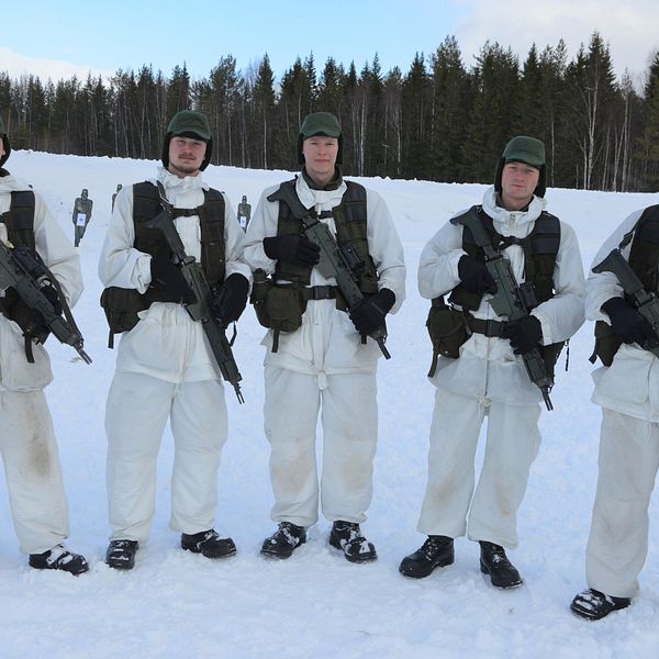 Fem unga militärer i vit vintermuntering står och håller i varsitt automatgevär.