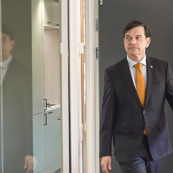 En man i kostym med orange slips går mot en dörr i en kontorsmiljö. Reflektionen av honom ses i det glasade elementet på vänster sida av bilden. Bakgrunden består av ljusa väggar och en dörr.