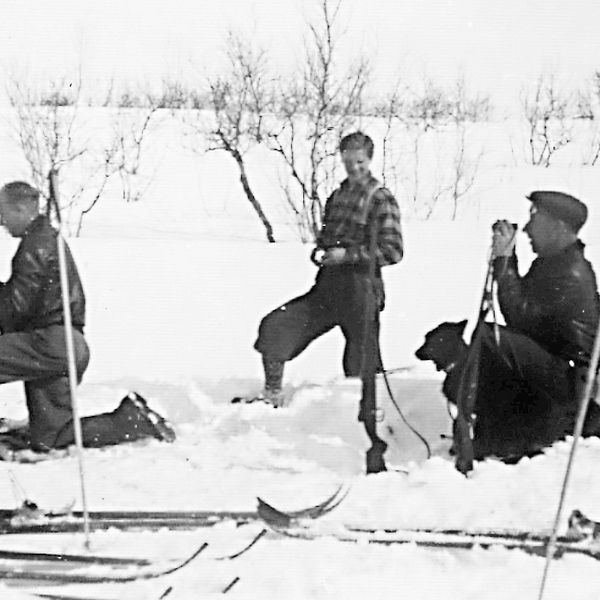 gammalt svartvitt foto på fyra män som sitter och står i snön, skidor bredvid, en håller gevär