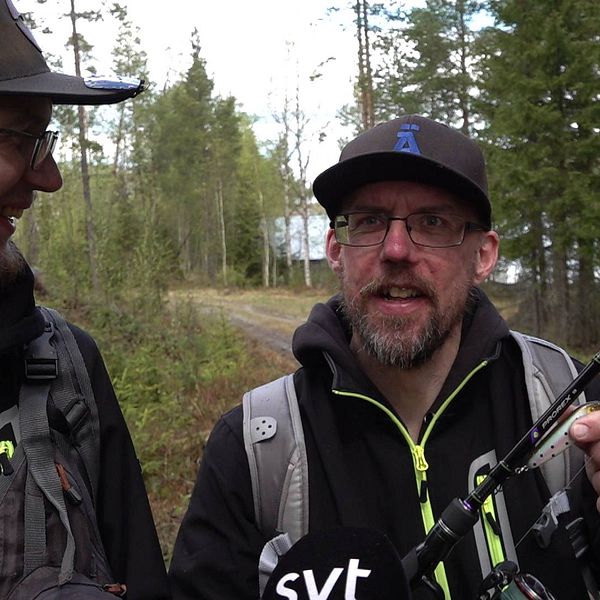 Två män med ett fiskespö står och flinar mot kameran på en skogsväg, sjö skymtar