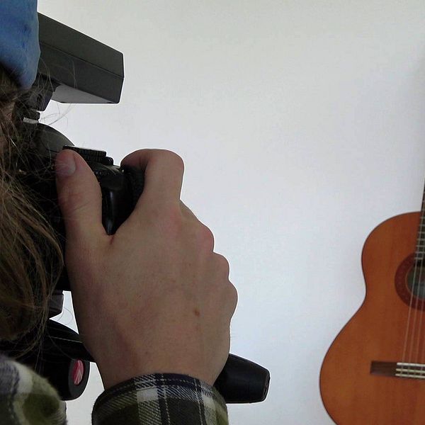 I bakgrunden ser vi en akustisk gitarr står mot en vit bakgrund. I förgrunden ser vi en person som har händerna på en kamera och fotograferar av gitarren.