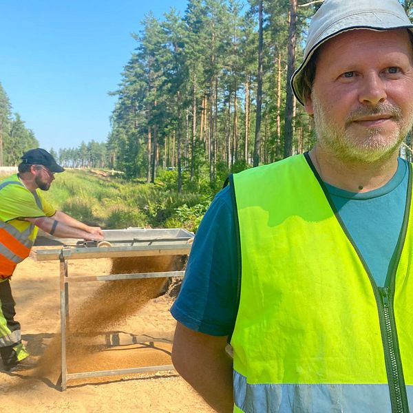 Bild på  Henrik Runeson, arkeolog och projektledare på Stiftelsen Kulturmiljövård. Han har på sig solhat och varselväst. I bakgrunden står hans kollega Anderas Bolin som sållar.