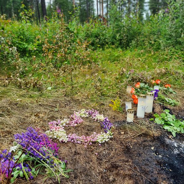 blonkrans och gravljus på marken vid en förkolnad fläck, skog bakom